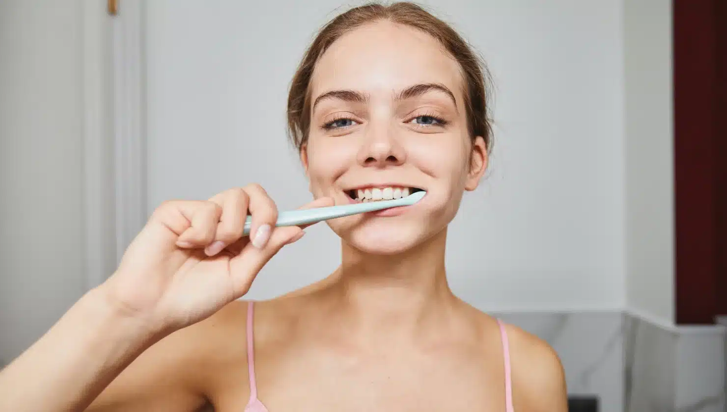Young woman brushing her teeth with a blue toothbrush, smiling in a bathroom setting, promoting dental hygiene and oral care.