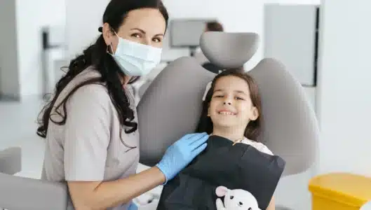 Pediatric dentist smiling with young girl in dental chair, promoting children's dental health and comfort during check-up.