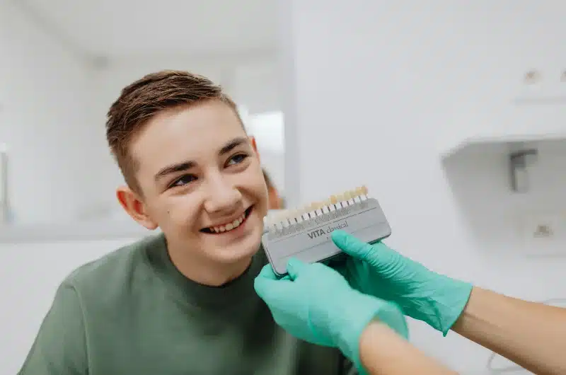 Teenager smiling while a dental professional compares teeth shade using a color guide in a pediatric dental office.