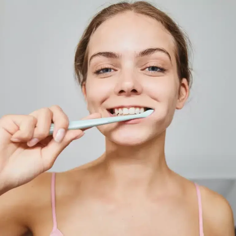 Smiling woman brushing her teeth with a light blue toothbrush, emphasizing oral hygiene and the importance of maintaining a healthy smile.